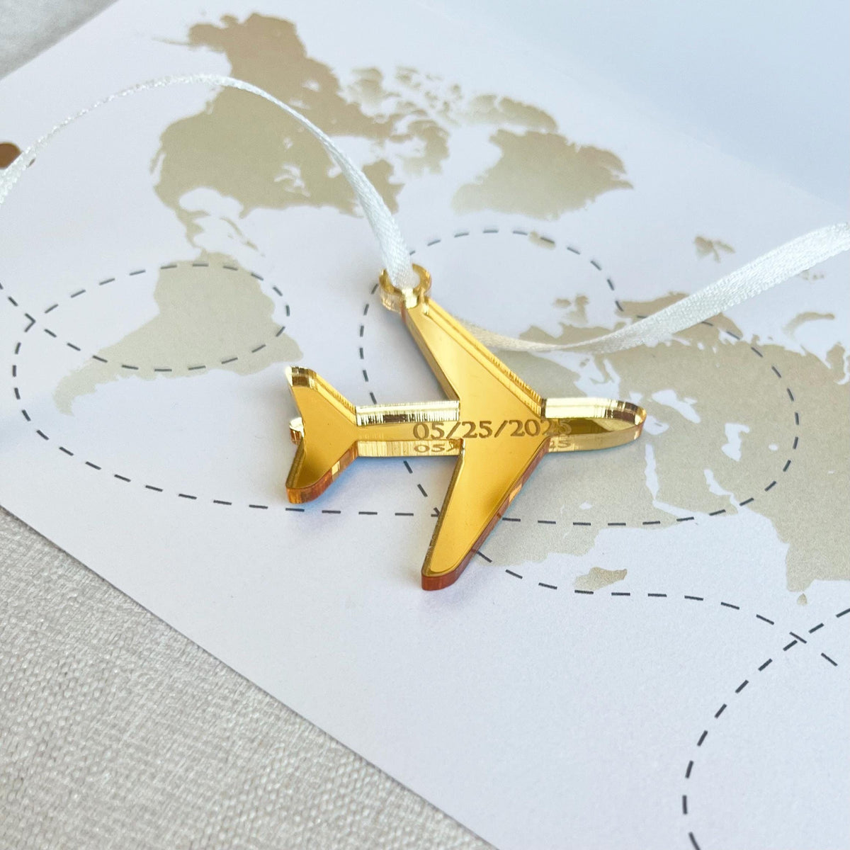 A unique airplane-shaped tag made from mirror gold acrylic with your wedding date elegantly laser engraved. The tag is laced with 3mm pearl satin ribbon for a touch of sophistication. It sits on a printed card with a world map and continents, creating a stunning travel-inspired presentation.