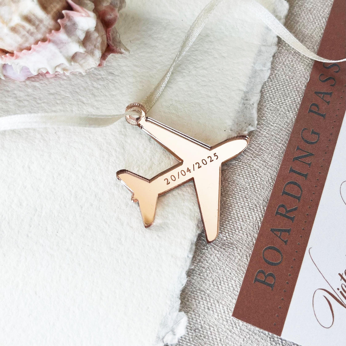 A unique airplane-shaped tag made from mirror rose gold acrylic, elegantly laser engraved with your wedding date. The tag is strung with a 3mm pearl satin ribbon for a touch of sophistication. It is placed on a textured card and a boarding pass is placed on the side - a passport-style invitation.