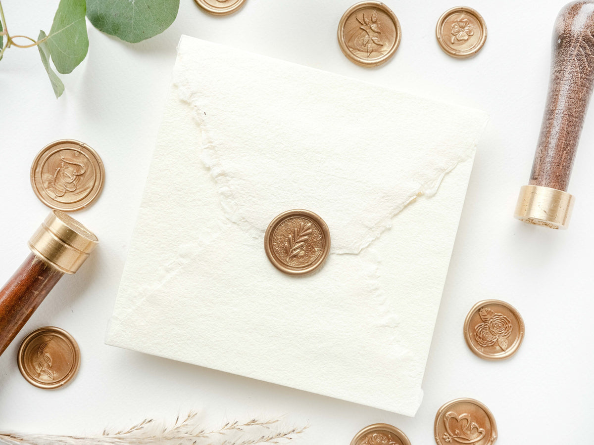 May include: A white envelope with a gold wax seal. The seal has a leaf design. There are other gold wax seals scattered around the envelope, including one with a flower design. There is a wooden handle with a gold base next to the envelope.